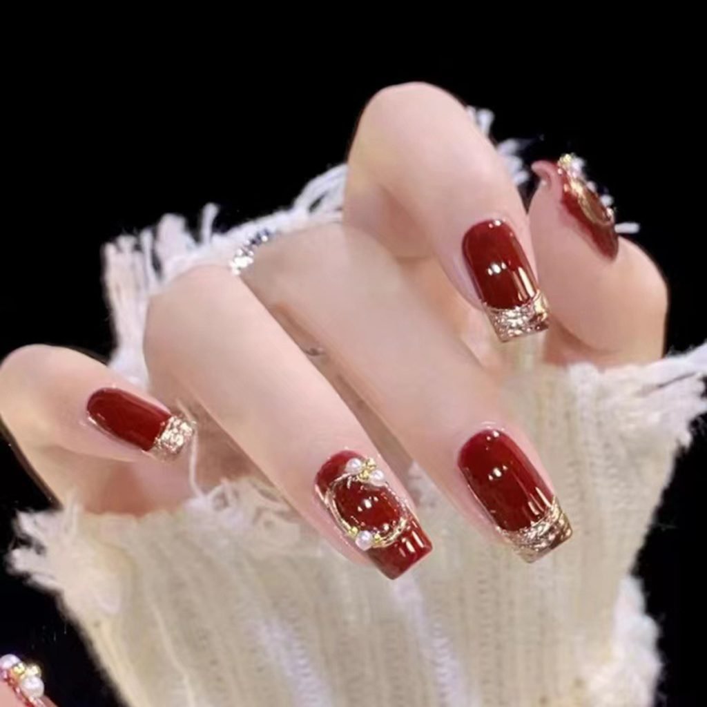 A hand showcasing a set of handmade squoval press-on nails in wine red with golden ribbon design, pearl accents, and a French tip.