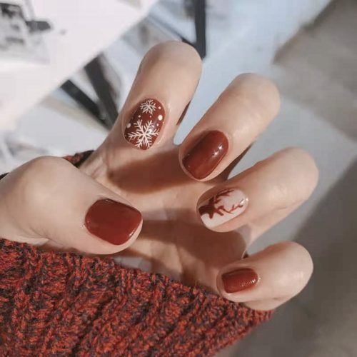 A hand modeling a set of squoval press-on nails in shades of wine red and light pink, featuring Christmas-themed reindeer and snowflake nail art.