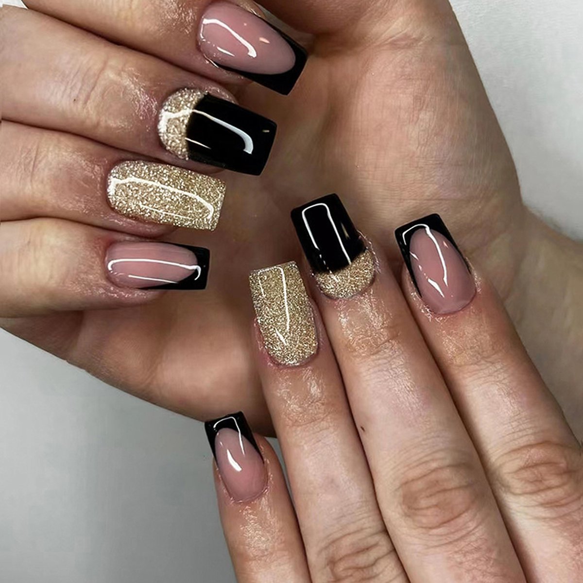 Hand wearing a variety of square-shaped fake nails in pink with black French tips, gold glitter, and black with gold glitter accents.