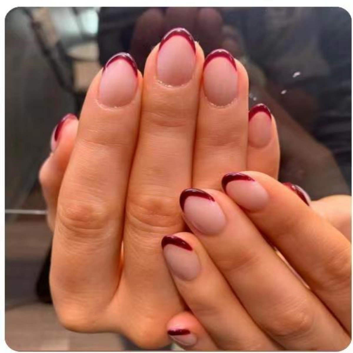 Two hands elegantly displaying short almond-shaped press-on nails, featuring a nude shade with vibrant red French tips