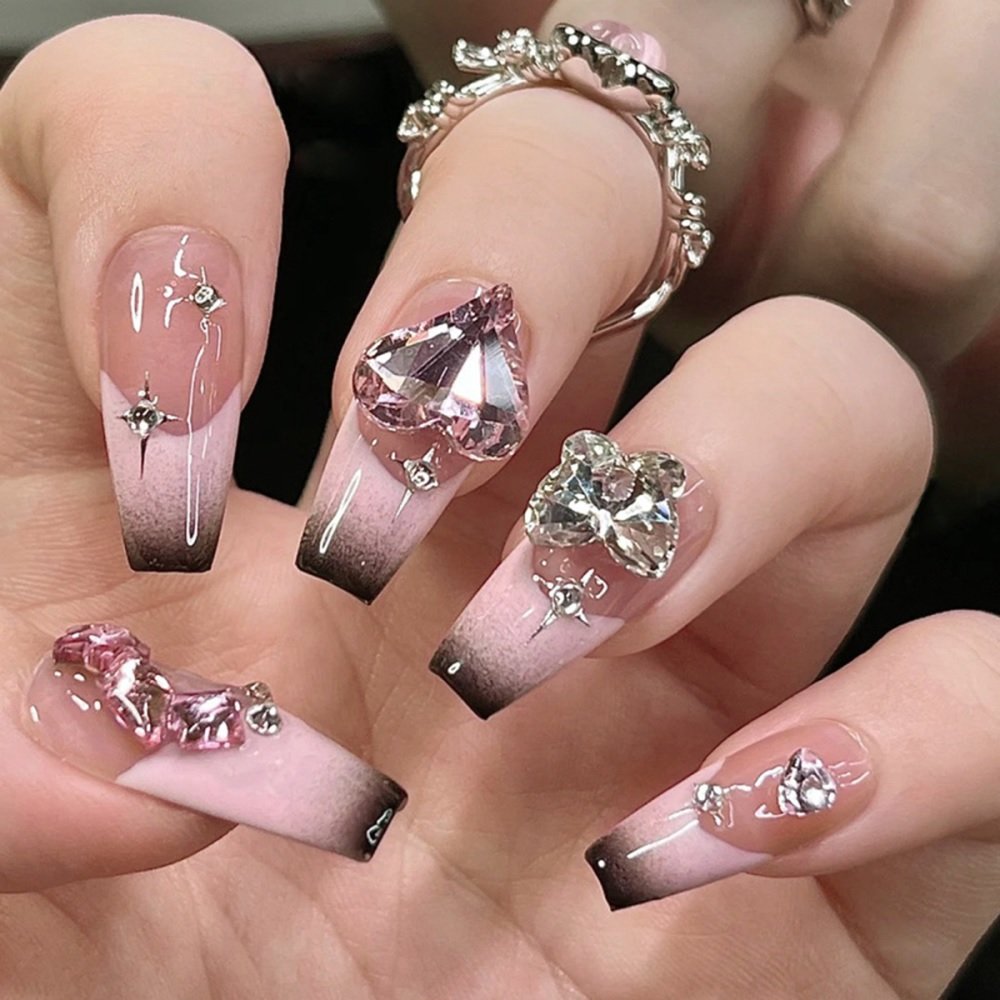 Hand showcasing coffin press-on nails in nude shade with French tips blending black and pink; accentuated with large diamond embellishments.